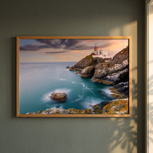 Baily Lighthouse Sunset, Howth Head framed print in a natural oak frame on a dark green wall beside a window, dappled plant shadow