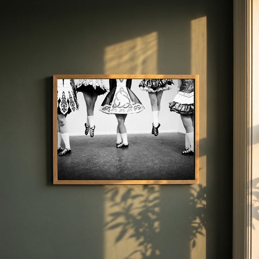 Irish Dancing Black & White framed print in a natural oak frame on a dark green wall beside a window, dappled plant shadow