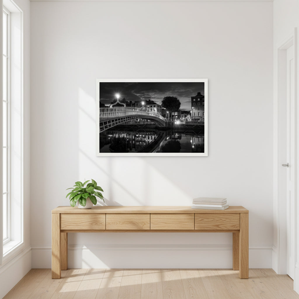 Black and white Ha’penny Bridge night photograph with city lights reflecting in the River Liffey, framed in a white wooden frame, 30x45 cm. Bright hallway with white walls and wooden console table, 30x45 cm Ha’penny Bridge black and white print in white frame hanging above, natural daylight, potted plant and books below, airy Dublin apartment vibe