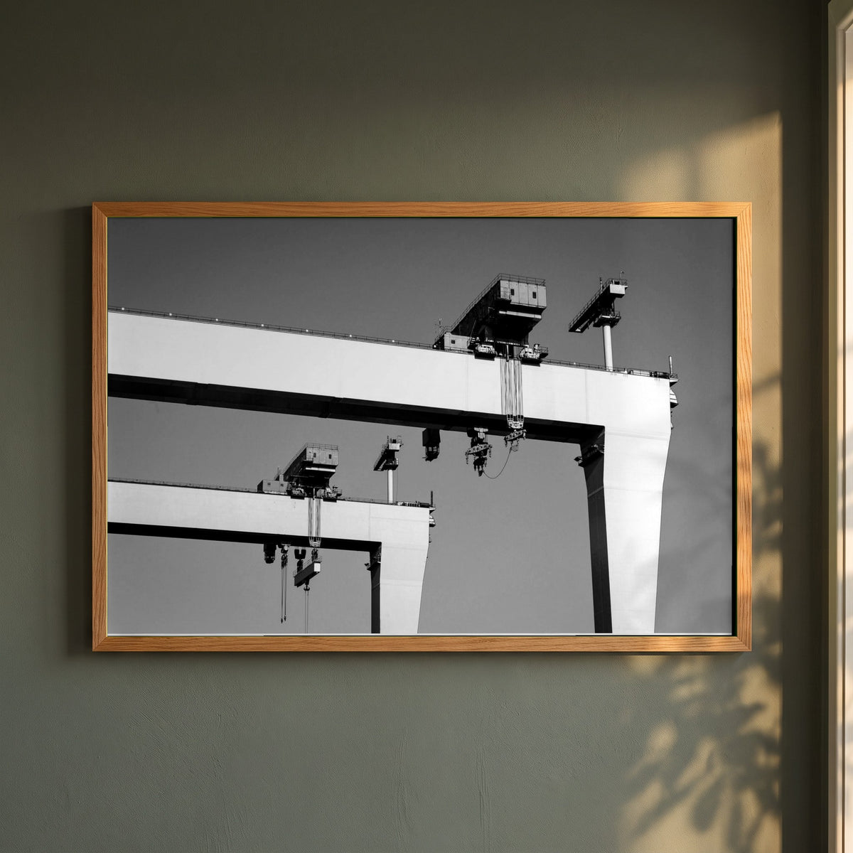 Harland & Wolff Cranes, Belfast framed print in a natural oak frame on a dark green wall beside a window, dappled plant shadow