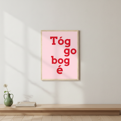 Tóg go bog é Irish language print in red on pink in a light oak frame above a low wooden shelf with a green vase