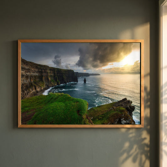 Cliffs of Moher framed print in a natural oak frame on a dark green wall beside a window, dappled plant shadow