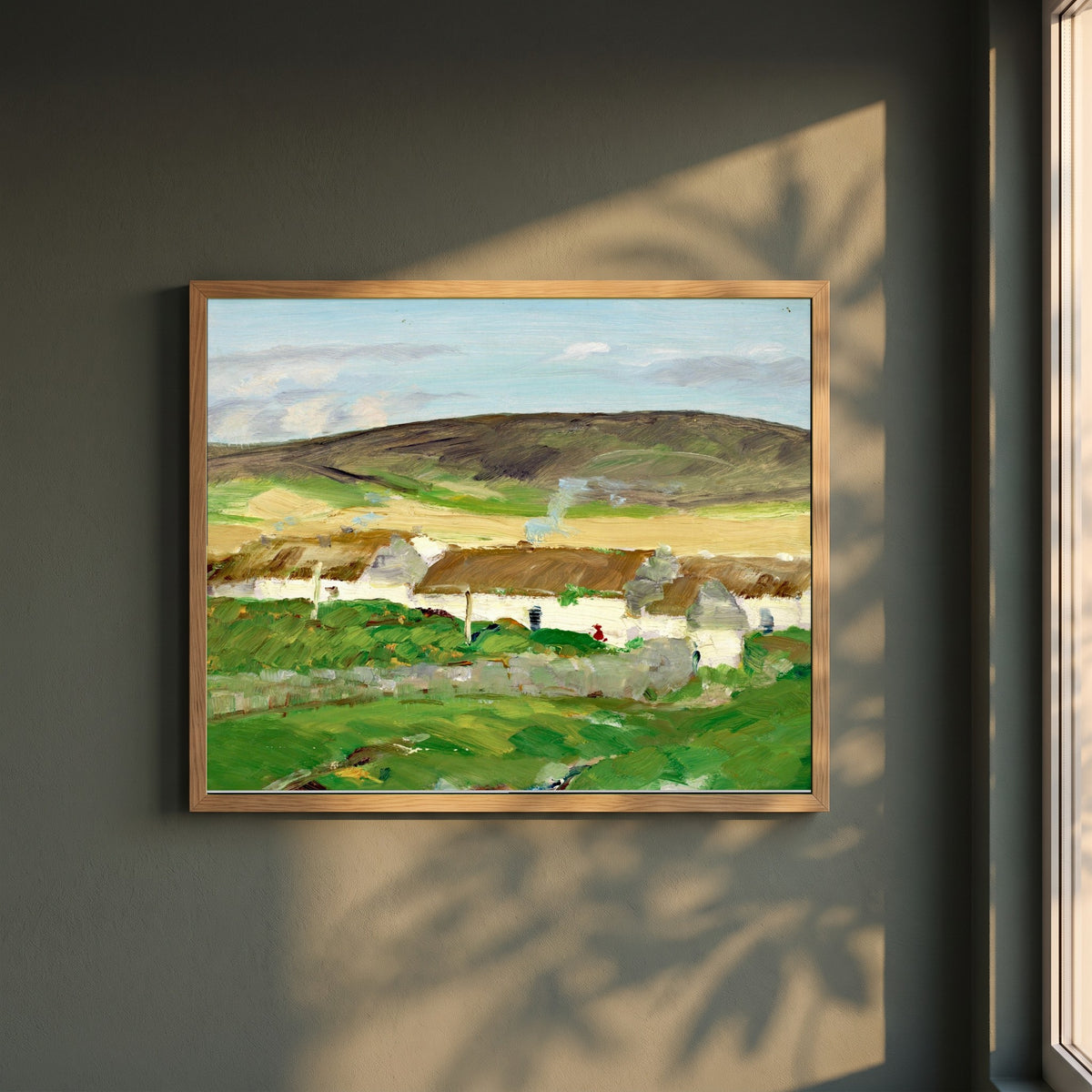 The White Cottage framed print in a natural oak frame on a dark green wall beside a window, dappled plant shadow